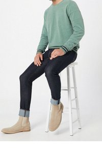 Sudadera verde con puños a rayas, jeans de mezclilla oscuros con puños doblados y botas de tobillo beige, llevadas mientras se está sentado en un taburete blanco.