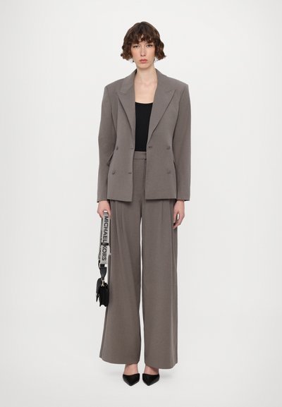 Grau doppelt geknöpfter Blazer über einem schwarzen Oberteil, kombiniert mit weit geschnittenen grauen Hosen. Schwarze spitze Pumps und eine Handtasche mit Logo-Tragegurt.
