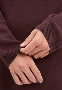 Bordowy sweter o miękkiej fakturze, z prążkowanymi mankietami. Ręce chwytają mankiet, pokazując szczegóły tkaniny i szwu.