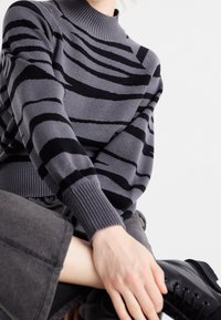 Pull gris à rayures zèbre noires, col montant côtelé et poignets côtelés, associé à un pantalon en tissu sombre et des bottes noires à lacets.