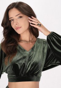 Top corto in velluto verde con scollo a V e maniche a puff. Collana in argento dorato e grande anello dorato completano il look. Texture liscia e morbida.