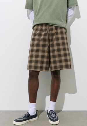 Homme portant un short marron à carreaux, des chaussettes blanches, des chaussures noires sans lacets avec des motifs blancs, un T-shirt vert par-dessus des manches longues blanches, debout contre un mur blanc.