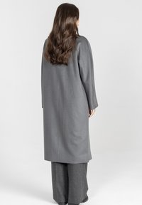 Długi szary płaszcz z teksturowanej tkaniny, z długimi rękawami, prostym krojem i długością do połowy łydki, zestawiony z szerokimi szarymi spodniami.