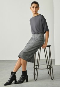 Frau, die auf einem Metallhocker sitzt und ein graues kurzärmliges Oberteil, einen karierter knielangen Rock und schwarze Absatzstiefel trägt, vor einem schlichten Hintergrund.