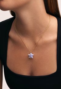 Collier en chaîne dorée présentant un pendentif en forme d'étoile de couleur violet clair avec des accents blancs et des motifs détaillés.