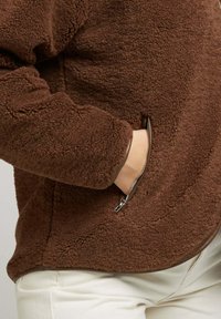 Pull en polaire marron avec une surface texturée, poches latérales zippées et un ourlet arrondi. Présente un design ajusté et des manches longues.
