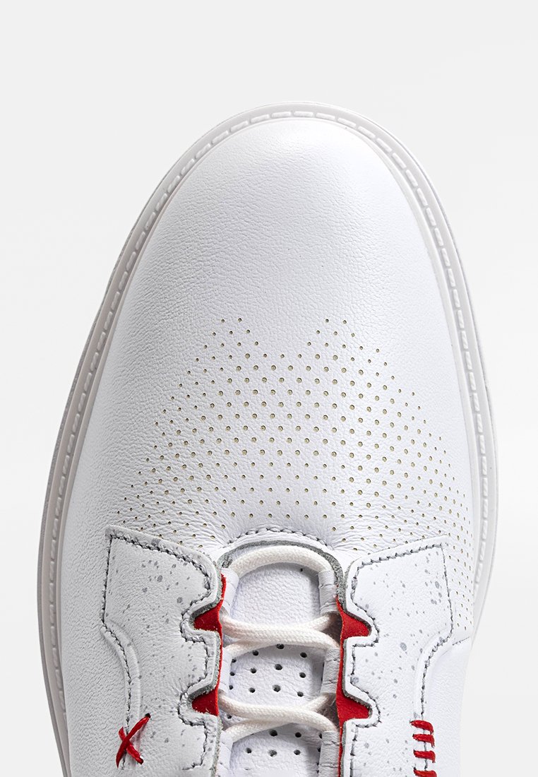 Chaussure en cuir blanc avec texture perforée, dotée d'accents et de lacets rouges. Bout rond et couture visible autour de la semelle.