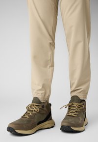 Bruine wandelschoenen van gestreept suede en mesh bovenkant, gecombineerd met beige, elastische onderbroek. Veters zijn lichtbruin en plat.