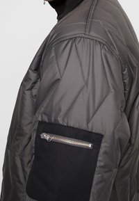 Grå pufferjakke med diamantquiltet tekstur, syede detaljer og en sort lynlåslomme på ærmet for ekstra opbevaring.
