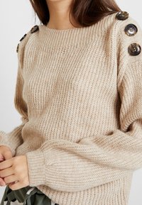 Vrouw in een beige gebreide trui met grote schildpadknopen op de schouders, handen losjes voor zich gevouwen, tegen een effen achtergrond.