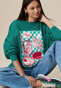 Zelená oversize mikina s potlačou jedla, okrúhly výstrih, kombinovaná s vysokým pásom svetlo modrých džínsy a zeleno-ružovými teniskami.