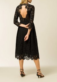 Svart spetsklänning med trekvartslängd ärmar, med en djup ryggskärning och scalloped fåll. Bärs med svarta sandaletter.