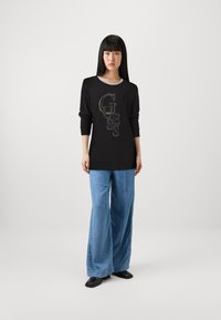 Maglietta nera a maniche lunghe con il logo "GUESS" in lettere dorate, abbinata a pantaloni in denim blu a gamba larga. Tessuto morbido, vestibilità comoda.