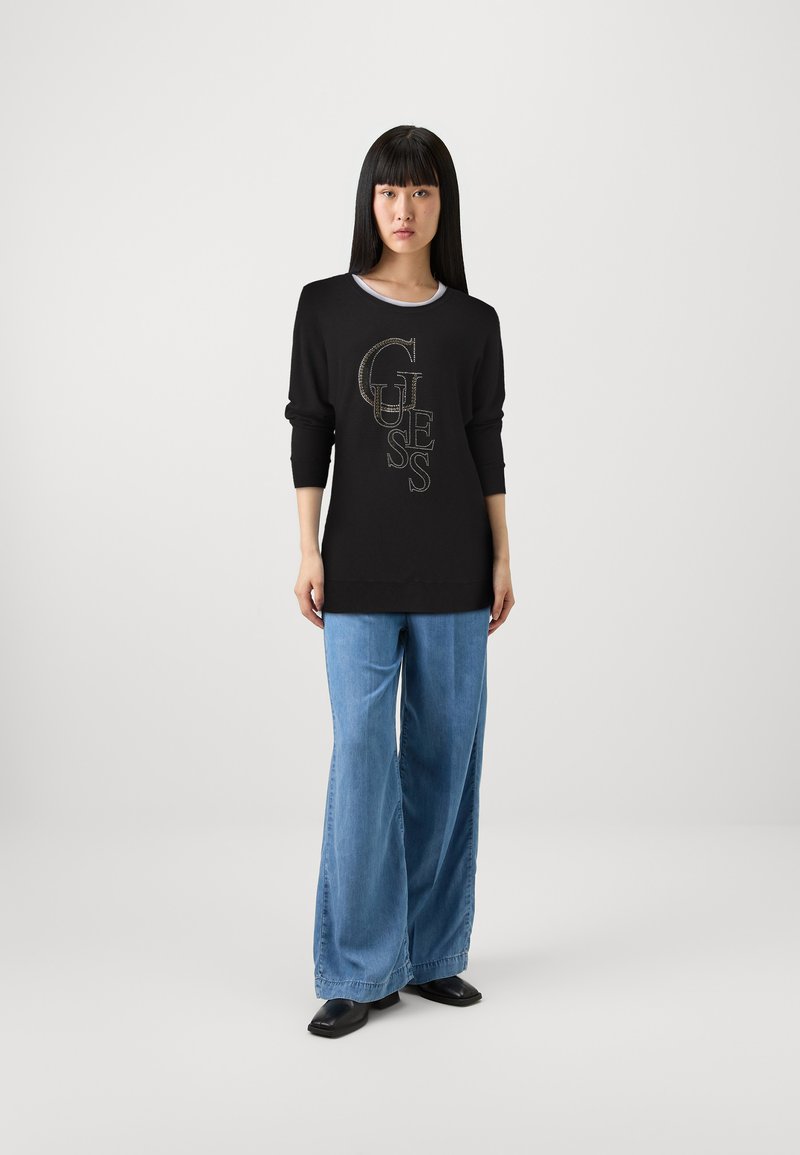 Maglietta nera a maniche lunghe con il logo "GUESS" in lettere dorate, abbinata a pantaloni in denim blu a gamba larga. Tessuto morbido, vestibilità comoda.