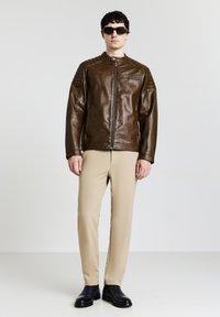 Uomo con giacca di pelle marrone scuro, pantaloni beige, scarpe nere e occhiali da sole neri in piedi contro uno sfondo bianco uniforme.