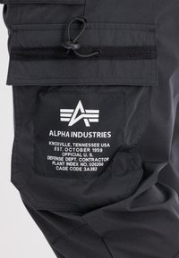 Närbild av svarta Alpha Industries byxor med en ficka, där vit logotyp och text redovisar företags- och kontraktsinformation.