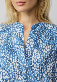 Blau-weiß gemusteter Stoff mit einem leichten, strukturierten Aussehen. Verfügt über ein V-Ausschnitt-Design und einen Knopfleistendetail.