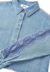 Camisa de mezclilla azul claro con cuello y botones al frente. Presenta un intrincado detalle de ganchillo azul claro a lo largo de una manga.