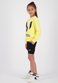 Felpa gialla a maniche lunghe con logo nero, abbinata a pantaloni cargo neri. Indossata con sneakers alte bianche con accenti neri e gialli.