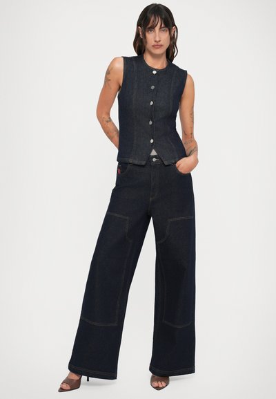 Mulher de pé com as mãos nos bolsos, vestindo um colete de ganga escuro sem mangas abotoado, jeans de perna larga em ganga escura e sandálias com salto castanhas.