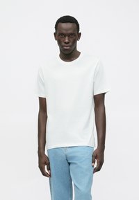 Giovane uomo che indossa una semplice maglietta bianca a maniche corte e jeans blu chiaro, in piedi contro uno sfondo bianco, con uno sguardo neutro.