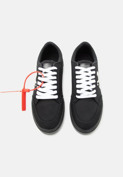 Baskets basses noires avec lacets blancs, logo latéral blanc et une étiquette en plastique rouge fixée au lacet de la chaussure gauche.