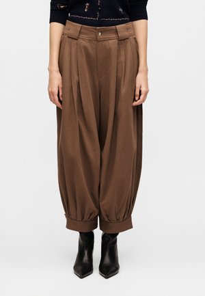 Brązowe spodnie z wysokim stanem, szerokimi nogawkami, plisami i podwiniętymi mankietami, noszone z czarnymi botkami z ostrym noskiem i ciemnym wzorzystym topem.