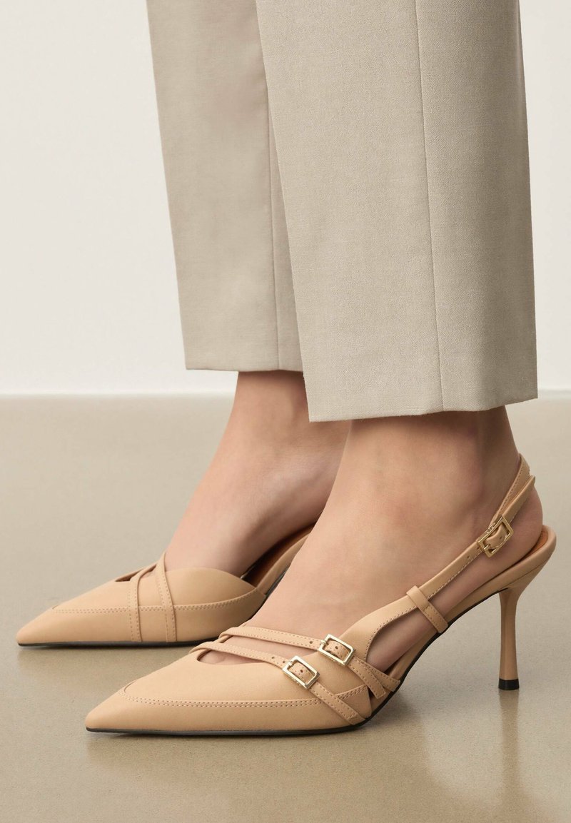 Escarpins beige à bride arrière avec bout pointu, dotés de brides croisées et de boucles dorées. Texture lisse et talons aiguilles fins.