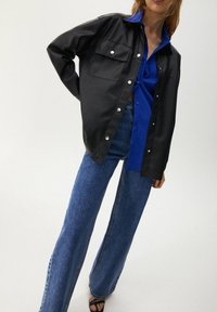 Kvinna som bär en svart knappjacka över en blå skjorta, tillsammans med vida blå jeans och svarta sandaler med öppen tå, står framför en enkel bakgrund.