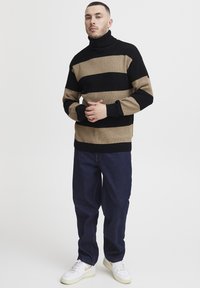 Stripet turtleneck genser i svart og tan ribbet tekstur, parret med mørkeblå jeans og hvite joggesko med gule detaljer.