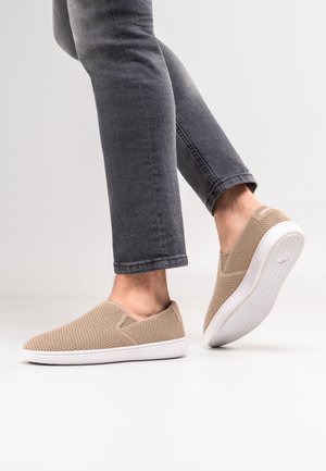 Personne portant des baskets beige à enfiler texturées et un jean gris foncé retroussé, un pied levé montrant la semelle de la chaussure.