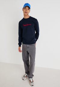 Sudadera navy con logo rojo de "Pepe Jeans", jeans grises y zapatillas grises. La gorra es azul; el conjunto tiene un corte casual y relajado.