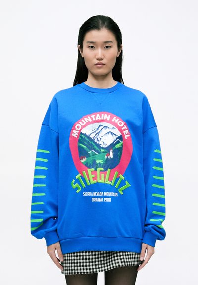 Sweatshirt bleu avec une coupe surdimensionnée, arborant un graphique circulaire de montagne en rouge et vert, et des accents rayés verts sur les manches. Le texte indique "Hôtel de Montagne Stieglitz."