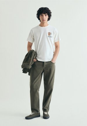 Jeune homme aux cheveux bouclés porte un t-shirt graphique crème, un pantalon vert olive et tient une veste assortie, debout les mains dans les poches.