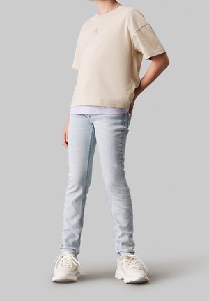 Béžové bavlněné oversized tričko s levandulovým logem, kombinované se světle modrými slim džínami a bílými teniskami na neutrálním pozadí.