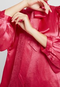 Blusa lucida di un profondo colore rosa con colletto a fiocco, maniche gonfie e polsini con bottoni. Presenta una texture liscia e una leggera lucentezza.