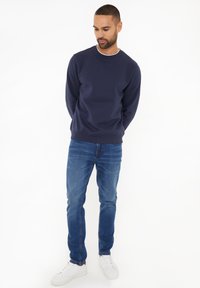 Granatowy sweter z okrągłym dekoltem, zestawiony z niebieskimi dżinsami i białymi butami sportowymi. Sweter ma gładką powierzchnię i ściągacze przy mankietach.