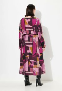 Langes, dunkelrosa Kleid mit geometrischen Mustern in Pink-, Lila- und Senfgelb-Tönen, langen Ärmeln und elastischem Bund, kombiniert mit schwarzen Stiefeln.