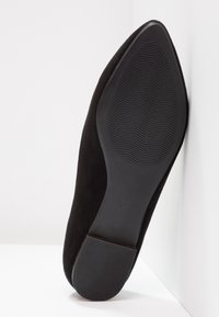 Semelle de chaussure noire à bout pointu avec des grips texturés au talon et à l'avant-pied, inclinée en position verticale sur un fond blanc.