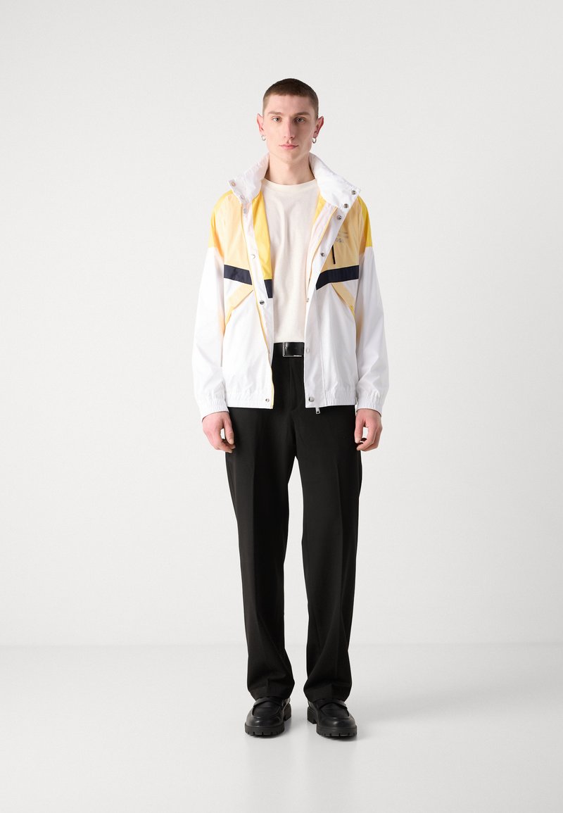 Veste légère pour homme en blanc avec des accents jaunes et marine, dotée d'un col montant, d'une fermeture éclair et de poignets élastiques. Associée à un pantalon noir.