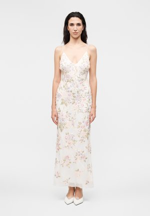 Ženska v beli obleki brez rokavov z rožnatim in zelenim cvetličnim vezenjem ter belimi visokimi petami s koničastim sprednjim delom, stoji pred enostavnim ozadjem.