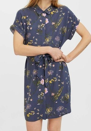 Femme portant une robe à manches courtes, bleue marine à fleurs, avec un col et une ceinture en tissu nouée à la taille.
