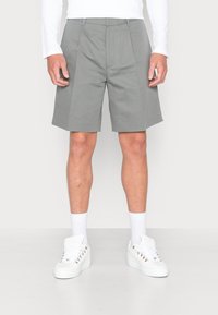 Pantaloni corti grigi chiari con pieghe, realizzati in tessuto morbido. Indossati con sneakers bianche e calze. Linee pulite e vestibilità moderna.