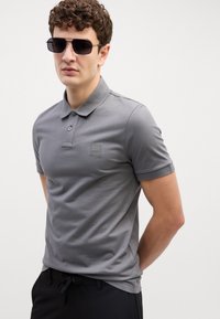 Šedé polo tričko vyrobené z bavlny, so stojatým golierom, zapínaním na dva gombíky a malou nášivkou s logom. Nosené s tmavými slnečnými okuliarmi a nohavicami.
