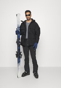 Jachetă neagră de ski cu multiple buzunare, cămașă gri, pantaloni negri de ski, mănuși albastre, ochelari de soare, ținând schiuri albastre și albe cu legături.