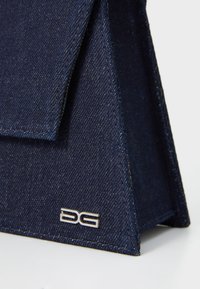 Mørkeblå denimhåndtaske med en trekantet form, der har et sølvlogo nederst til højre hjørne og en foldet låseklap.
