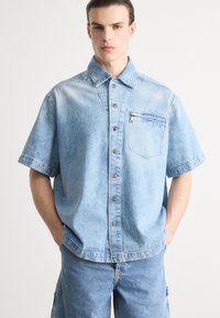 Camisa de mezclilla azul claro de manga corta con botones en la parte delantera, cuello puntiagudo, bolsillo en el pecho y corte relajado con costuras tonales.