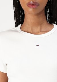 T-shirt bianca a coste con maniche corte e scollatura rotonda, con un piccolo logo ricamato in rosso, bianco e blu sul petto.