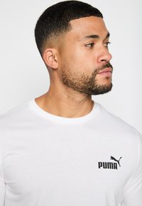 Camiseta de algodón blanca con un logo negro de Puma en el lado izquierdo del pecho. El cuello redondo tiene una textura acanalada. Diseño suave y ajustado.