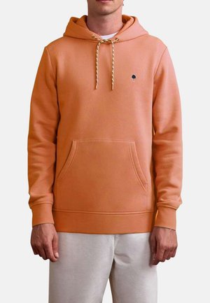 Homme portant un sweat à capuche orange clair avec poche avant et cordons, associé à un pantalon de couleur claire, sur un fond uni.